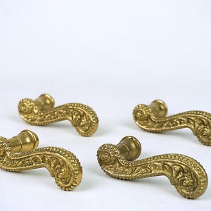 Puede incluir: Cuatro manijas de puerta antiguas de latón con diseños florales ornamentados. Las manijas son curvas con una superficie detallada y texturizada y un tono dorado cálido. Están dispuestas sobre un fondo blanco, mostrando su intrincada artesanía.