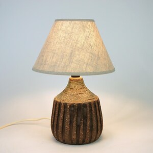 May include: A decorative table lamp with a textured, brown ceramic base and a light gray lampshade. The base has vertical grooves and a lighter-colored top section. The lamp is designed for indoor use and has a neutral color palette.