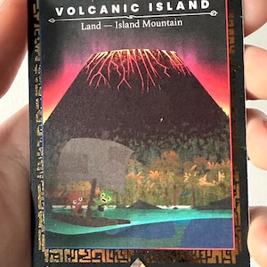 May include: A card depicting a volcanic island. The image shows a dark, cone-shaped mountain with red and orange lava flows. The text "VOLCANIC ISLAND" is at the top, with "Land - Island Mountain" below. A boat with cartoon characters is on the water.