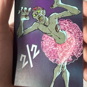 May include: A card featuring a cartoon zombie wearing a pink tutu and a crown. The zombie has green skin, yellow eyes, and an open mouth. The card has the text "2/2" in the lower left corner.