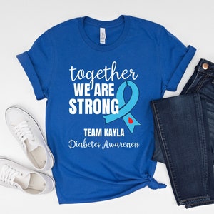 Op de afbeelding: Koningsblauw t-shirt met witte tekst die luidt "together WE ARE STRONG" en "TEAM KAYLA Diabetes Awareness". Een lichtblauw lint met een rode druppelafbeelding is ook te zien. Het shirt wordt gecombineerd met een blauwe jeans en witte sneakers.