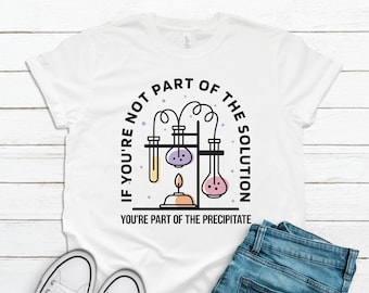 May include: A white t-shirt featuring a black graphic of laboratory glassware, including test tubes and a Bunsen burner. The text reads, "IF YOU'RE NOT PART OF THE SOLUTION YOU'RE PART OF THE PRECIPITATE," with purple and yellow accents.