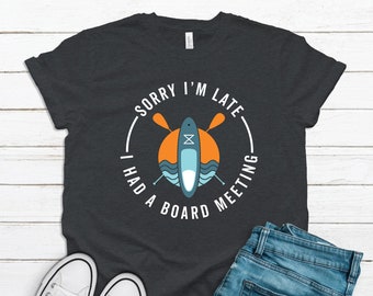 Op de afbeelding: Een donkergrijs T-shirt met korte mouwen en een afbeelding van een paddleboard en twee gekruiste peddels voor een oranje zon en blauwe golven. De tekst SORRY I'M LATE I HAD A BOARD MEETING staat in witte hoofdletters in een cirkel.