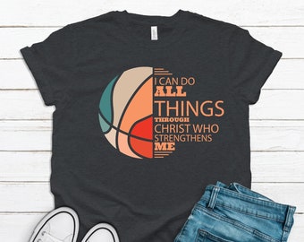 May include: Dark gray t-shirt with a basketball graphic in teal, tan, orange, and red. The shirt features the text "I CAN DO ALL THINGS THROUGH CHRIST WHO STRENGTHENS ME." White sneakers and blue jeans are also visible.