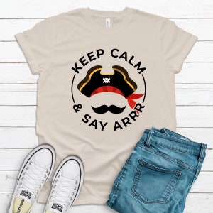 May include: A beige t-shirt with a black circle graphic that says "Keep Calm & Say Arrr". The graphic includes a pirate hat with a skull and crossbones and a black mustache.