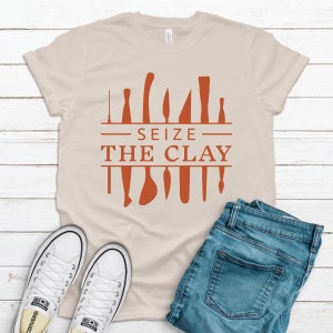 Op de afbeelding: Een beige T-shirt met een bruine afbeelding van pottenbakkersgereedschap en de tekst "Seize the Clay".