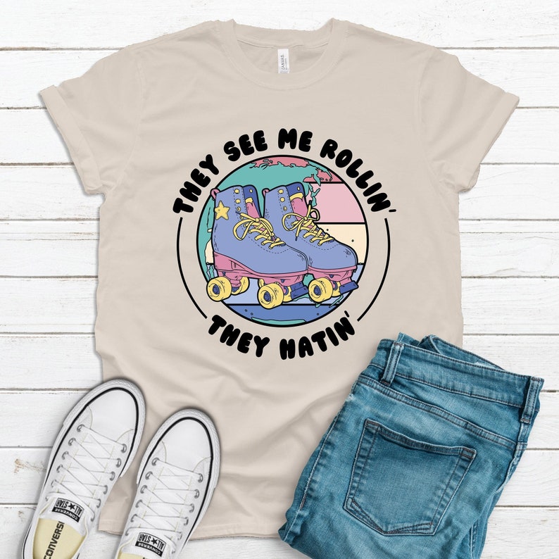 Natural colored graphic t-shirt with retro roller skate design that says They See Me Rollin They Hatin