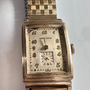 May include: A vintage gold-toned Longines wristwatch with a rectangular face. The watch features a cream-colored dial with black numerals and hands. The watch band is a gold-toned metal link style. The watch face reads "Longines" and "Swiss".
