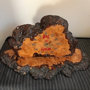 May include: A wooden clock with a natural, rustic design. The clock face is made of light brown wood with a visible grain, and the text "God is Love" is written in red. The clock hands are black.