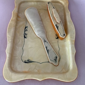 May include: A vintage, cream-colored plastic tray with a decorative, scalloped edge. The tray features a white plastic shoehorn and a small, rectangular, cream-colored box with a brown lid.