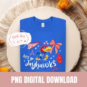 Op de afbeelding: Een koningsblauw t-shirt met een grafisch ontwerp met de tekst "Let's Go Jayhawks" en een cartoon Jayhawk-mascotte. Het ontwerp bevat een rode strik, een foamvinger en de tekst "KU". De woorden "PNG DIGITAL DOWNLOAD" staan onderaan.