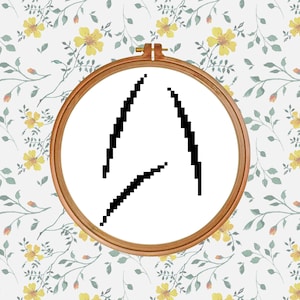 May include: A wooden embroidery hoop contains a white fabric with a black pixelated design. The design shows three angled lines. The background is a floral pattern with yellow flowers and green leaves.