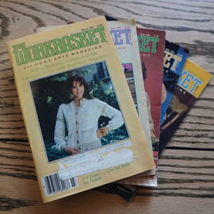 May include: A stack of vintage "Workbasket" magazines with colorful covers. The March 1984 issue features a woman wearing a cardigan. Other issues are visible, showcasing various cover designs and text.