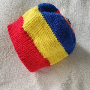 May include: A knitted beanie hat with red, yellow, and blue stripes. The hat is made from a soft, textured yarn and has a folded brim. The colours are bright and the hat appears to be handmade.
