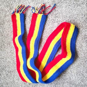 May include: A handmade crochet scarf in a vibrant color scheme of red, yellow, and blue. The scarf features a striped pattern with each color forming a distinct band. The ends of the scarf have fringe in matching colors.