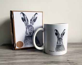 Può includere: Una tazza in ceramica bianca con un'illustrazione in bianco e nero di una lepre. La tazza è appoggiata su una superficie di legno accanto a una scatola con un'illustrazione simile di una lepre.