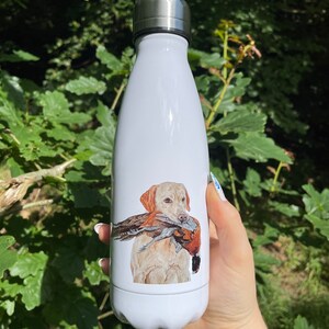 Pu&ograve; includere: Borraccia isolata bianca con tappo argentato. La borraccia presenta un'illustrazione dettagliata di un Labrador Retriever giallo che tiene un fagiano. Lo sfondo &egrave; verde naturale con la luce del sole.