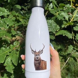 May include: White insulated water bottle with a stag illustration. The bottle has a silver cap and is held against green foliage. The stag illustration is in shades of brown and tan. The bottle is designed to keep drinks cold.