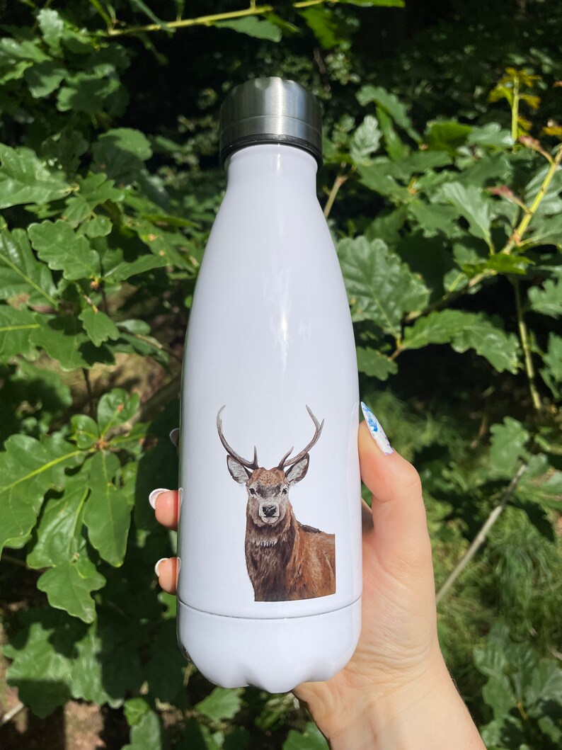 Pu&ograve; includere: Borraccia termica bianca con illustrazione di un cervo. Il tappo &egrave; argentato e la borraccia &egrave; tenuta contro un fogliame verde. L'illustrazione del cervo &egrave; nei toni del marrone e del beige.