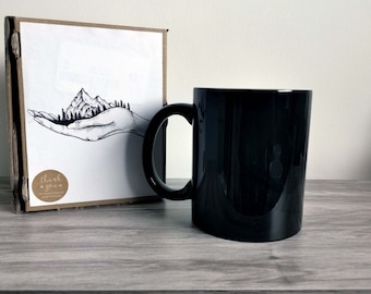 Può includere: Tazza in ceramica nera con una mano bianca che tiene una catena montuosa e alberi. La tazza è accanto a una scatola con un biglietto di ringraziamento.