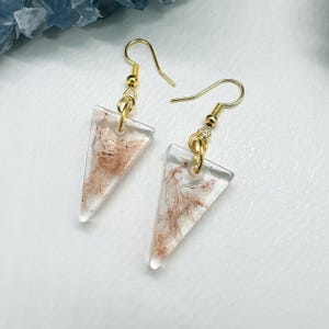 May include: A pair of clear, triangular earrings with gold-colored hooks. The earrings have a marbled, reddish-brown design within the clear resin. The earrings are displayed on a white surface.