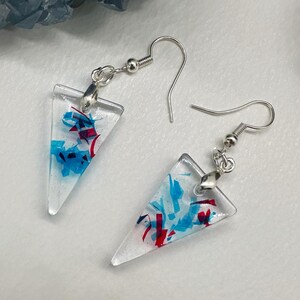 May include: A pair of clear, triangular earrings with silver-colored hooks. The earrings are filled with blue and red confetti-like pieces. The earrings are displayed on a white surface.
