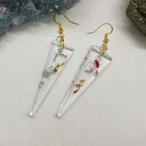 May include: A pair of clear resin earrings with gold-colored fish hooks. The earrings are triangular and contain silver, gold, and red flakes. The earrings are displayed on a white surface.
