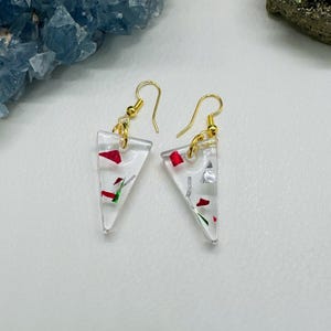 May include: A pair of clear, triangular earrings with gold-colored hooks. The earrings are filled with red, silver, and green confetti-like pieces. The earrings are displayed on a white surface.