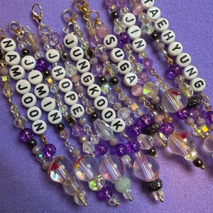 Puede incluir: Colección de llaveros de cuentas en tonos morados, transparentes y dorados. Cada llavero presenta un nombre deletreado con cuentas de letras blancas. Los llaveros están adornados con varias cuentas y dijes. El texto "with ARMY" es visible.