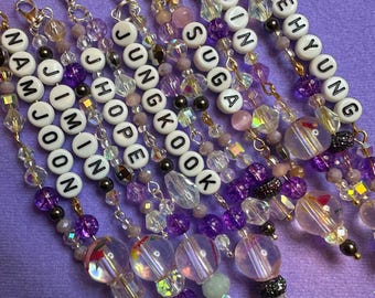 May include: A collection of beaded keychains in shades of purple, clear, and gold. Each keychain features a name spelled out with white letter beads. The keychains are adorned with various beads and charms. The text "with ARMY" is visible.