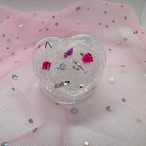 May include: A clear, heart-shaped container filled with silver glitter and colorful confetti. The container sits on a pink, sparkly fabric. The confetti includes pink, purple, and silver shapes.