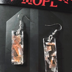 May include: A pair of rectangular earrings with silver hooks. The earrings are clear with embedded copper and silver-colored fragments. A black ribbon with the text "J-HOPE" in red is visible in the background.