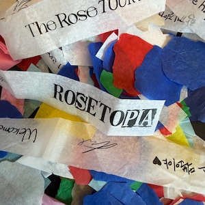 Puede incluir: Una colección de confeti de papel de colores en tonos rojo, azul, verde y amarillo, mezclado con tiras de papel blanco impresas con texto negro que incluye The Rose TOUR 2026, ROSETOPIA, Welcome, además de firmas manuscritas y escritura coreana.