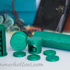 Puede incluir: Una colección de objetos verdes impresos en 3D, que incluyen un recipiente cilíndrico con el texto "CharmCaster", varias piezas redondas y una estructura alta y ranurada. Los objetos están dispuestos sobre una superficie blanca, con objetos borrosos turquesa y marrón al fondo.