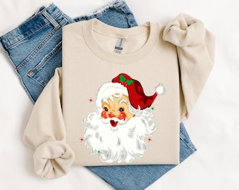 Op de afbeelding: Een beige sweatshirt met een vintage-stijl illustratie van het gezicht van Sinterklaas. Sinterklaas draagt een rode muts met een groene hulsttak en witte rand. Zijn baard is wit en zijn ogen zijn blauw.