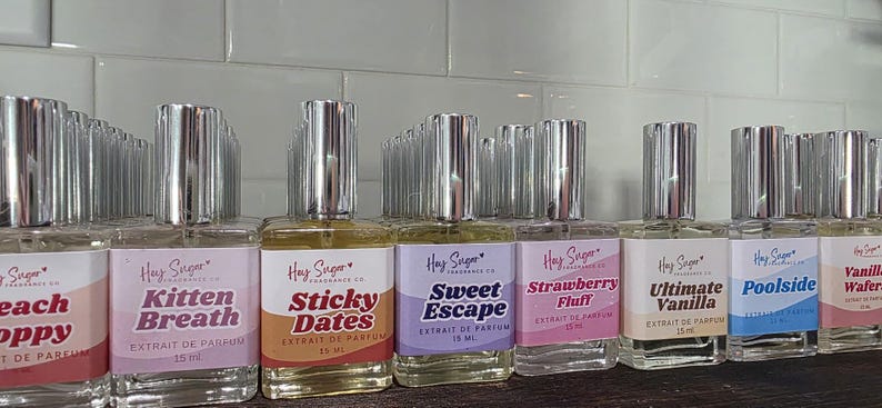 May include: A collection of Hay Sugar Fragrance Co. perfume bottles with silver caps. Each bottle has a label with a different scent name, including "Kitten Breath," "Sticky Dates," and "Sweet Escape." The bottles are arranged on a shelf.