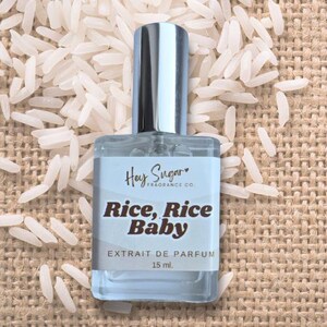 以下が含まれることがあります： Hey Sugar Fragrance Co.の「Rice, Rice Baby」とラベルされた、銀色のキャップが付いた透明な香水瓶。香水瓶には15mlのエクストレドパルファムが入っています。背景には、ジュートの表面に散らばる白い米粒が写っています。