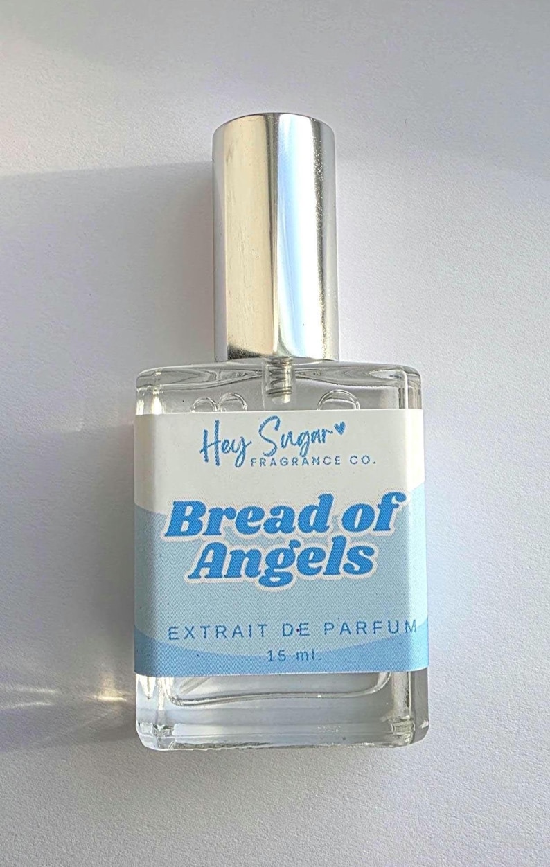 May include: A clear glass perfume bottle with a silver cap. The label reads "Hey Sugar Fragrance Co." and "Bread of Angels" in blue text. The bottle contains 15 ml of perfume.