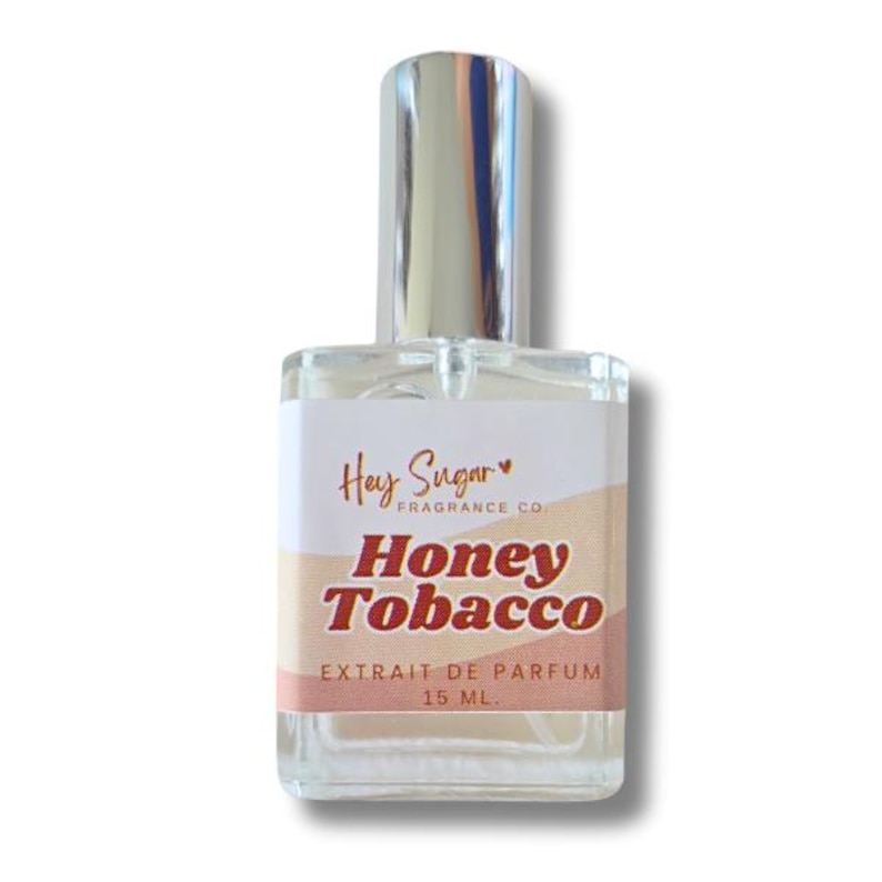 May include: A clear glass perfume bottle with a silver cap. The label reads "Hey Sugar FRAGRANCE CO." and "Honey Tobacco." The bottom of the label says "EXTRAIT DE PARFUM 15 ml."