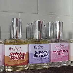 以下が含まれることがあります： 銀色のキャップが付いたHay Sugar Fragrance Co.の香水ボトルのコレクション。各ボトルには、「Kitten Breath」、「Sticky Dates」、「Sweet Escape」など、さまざまな香りの名前が記載されたラベルが付いています。ボトルは棚に並べられています。