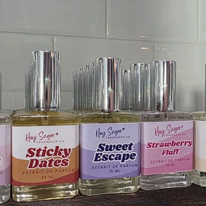 May include: A collection of Hay Sugar Fragrance Co. perfume bottles with silver caps. Each bottle has a label with a different scent name, including "Kitten Breath," "Sticky Dates," and "Sweet Escape." The bottles are arranged on a shelf.