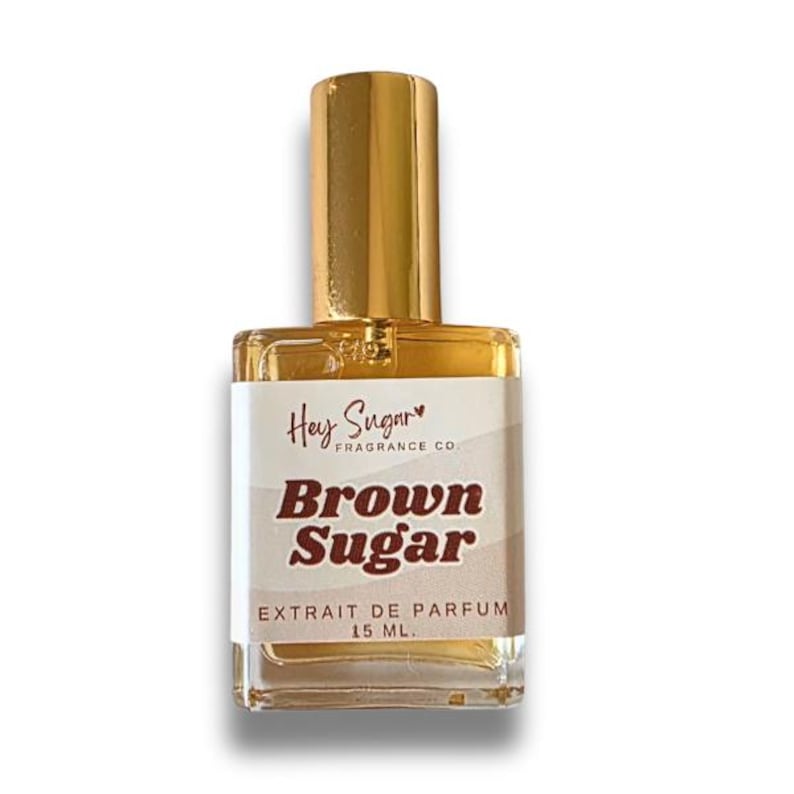 Puede incluir: Botella de perfume rectangular de vidrio con tap&oacute;n dorado. La etiqueta dice "Hey Sugar" y "Brown Sugar" con "Extrait de Parfum 15 ML." El l&iacute;quido interior es de color &aacute;mbar. La botella est&aacute; sobre un fondo blanco.