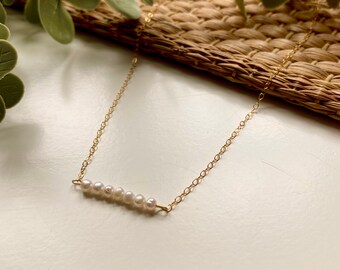 May include: Delicate gold necklace featuring a bar pendant adorned with seven small, white pearls. The chain is a fine, linked design. The necklace is displayed on a neutral background, highlighting its elegant simplicity.