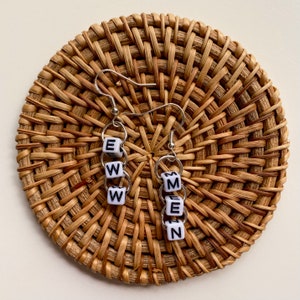 May include: Silver-tone drop earrings spelling "EWW" and "MEN" with white and black lettered beads. The earrings are suspended from silver hoops and hooks. The earrings are displayed on a round, woven, light brown placemat, similar to a coaster.