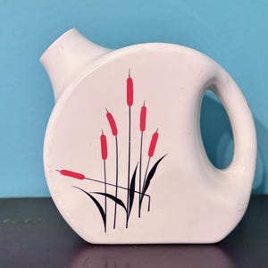 May include: A white ceramic pitcher with a rounded body and a handle. The pitcher features a design of red cattails and black grass. The spout is angled, and the pitcher is set against a blue background.