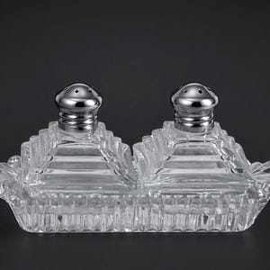 May include: A clear glass condiment set featuring two square, stepped-pyramid containers with silver-toned metal lids, each with small holes, resting on a rectangular, ribbed glass tray with decorative handles on both ends.