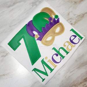 May include: Green, purple, and gold embroidered design of a jester's hat and mask with the number 7 and the name Michael.