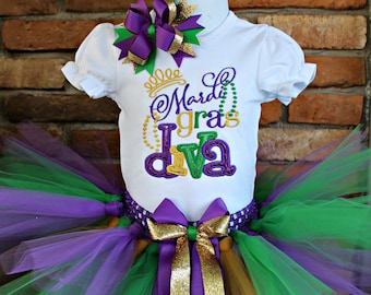 May include: A white shirt with the text "Mardi Gras Diva" in purple, gold, and green lettering. The shirt is paired with a green, purple, and gold tulle tutu with a purple and gold bow.