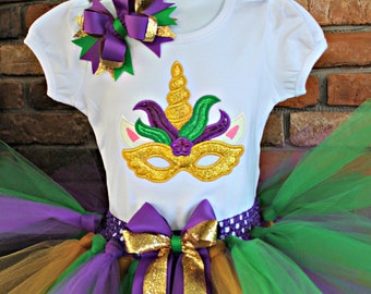 May include: A white t-shirt with a gold, green, and purple unicorn mask design. The shirt is paired with a multi-layered tulle skirt in gold, green, and purple. The skirt has a purple ribbon with gold glitter accents tied around the waist.