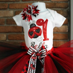 May include: A red and black tulle tutu with a red and black ladybug print ribbon. A white onesie with a red ladybug and the number one on it. The onesie also has the name Bella on it. The onesie is topped with a red, black, and white bow.